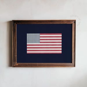 Könnte beinhalten: Gerahmter Druck der amerikanischen Flagge. Die Flagge zeigt rot-weiße Streifen mit einem blauen Kanton mit weißen Sternen. Der Druck ist vor einem dunkelblauen Hintergrund platziert und in einem dunklen Holzrahmen gerahmt.