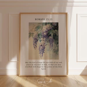 Op de afbeelding: Een ingelijste print met een aquarel van paarse blauwe regen bloemen die aan een tak hangen. De print bevat de tekst "Romeinen 15:13" en een citaat over hoop, vreugde en vrede.