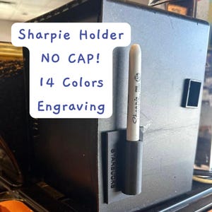 Può includere: Un porta Sharpie nero con un pennarello Sharpie bianco. Il supporto è attaccato a una superficie grigio scuro. Il testo sul supporto dice "Starbucks". Il testo sull'immagine dice "Sharpie Holder, NO CAP!, 14 Colors, Engraving."