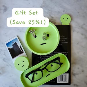Könnte beinhalten: Ein hellgrünes Geschenkset mit minimalistischem Gesichtsdesign, einschließlich einer Ablage, einem Brillenetui und einem Lesezeichen. Das Set enthält auch ein Foto und Schmuck. Der Text "Gift Set (Save 25% !)" wird angezeigt.