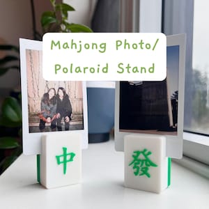 May include: White and green Mahjong tile photo stands holding Polaroid photos. The stands have Chinese characters in green. The text "Mahjong Photo/Polaroid Stand" is in a white box above the photos.