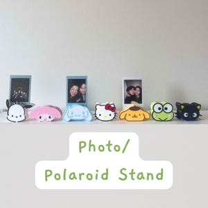 May include: A collection of colorful character-shaped photo and Polaroid stands. The stands are designed to hold small photos and feature popular cartoon characters in various colors, including white, pink, blue, yellow, green, and black. The text "Photo/Polaroid Stand" is visible.