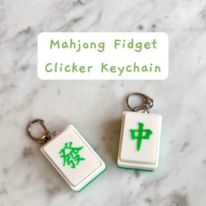 May include: Two white and green Mahjong Fidget Clicker Keychains with silver key rings. One keychain has a green Chinese character, and the other has a different green Chinese character. The keychains are on a marble surface.