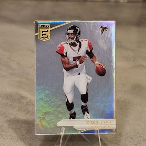 May include: A Michael Vick football trading card from the Elite series. The card features a player in a white and red uniform, holding a football. The card is displayed on a clear stand.