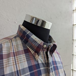 May include: A button-down shirt with a plaid pattern in shades of blue, burgundy, tan, and white. The shirt is displayed on a black mannequin head with a silver collar. The shirt has a button-down collar and a chest pocket.