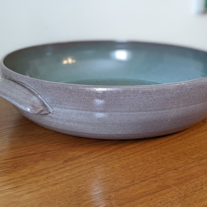 Könnte beinhalten: Eine große, hellgraue Servierschüssel aus Keramik mit einer hellblauen Glasur im Inneren. Die Schüssel hat eine abgerundete Form und einen einzigen Griff an einer Seite.