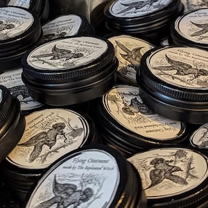 May include: A collection of small, black metal tins with white labels. Each label features a detailed illustration of a bird in flight and the text "Flying Ointment made by The Replenished Witch."