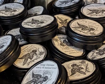 Pode incluir: Uma coleção de pequenas latas de metal preto com rótulos brancos. Cada rótulo apresenta uma ilustração detalhada de um pássaro em voo e o texto "Flying Ointment made by The Replenished Witch". As latas são mostradas em close-up.