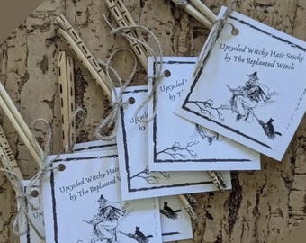 Pode incluir: Vários pacotes de paus de cabelo de bruxa reciclados são exibidos, cada um amarrado com barbante e acompanhado por uma etiqueta. As etiquetas apresentam uma ilustração em preto e branco de uma bruxa em uma vassoura, com o texto "Upcycled Witchy Hair Sticks by The Replanted Witch."