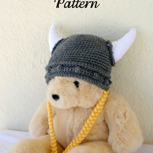 May include: A gray crocheted Viking helmet with white horns, worn by a beige teddy bear. The bear is wearing a yellow braided strap around its neck.