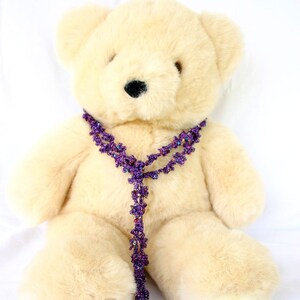 May include: A cream-colored teddy bear wearing a purple beaded necklace. The necklace is made of small, flower-shaped beads.