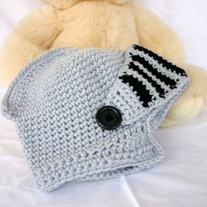 May include: A gray crocheted baby hat with a black and white striped brim and a black button. The hat is lying on a white surface.