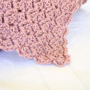May include: A close-up of a crocheted blanket in a dusty rose color. The blanket has a delicate, lacy pattern and is made with soft yarn.