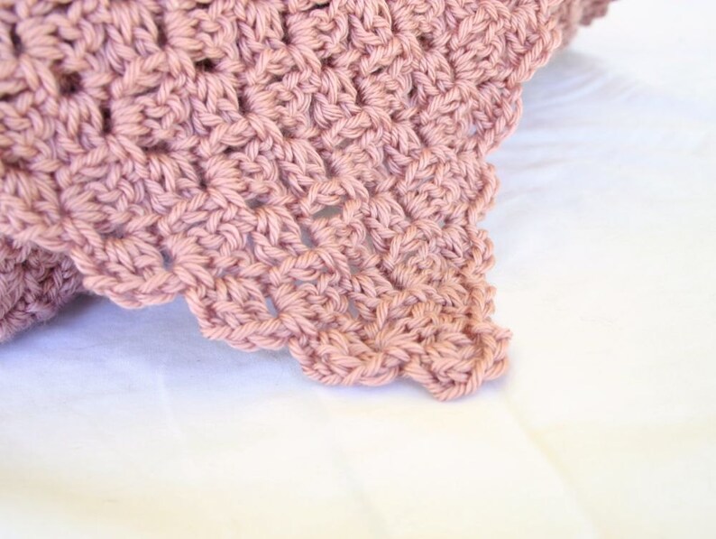 May include: A close-up of a crocheted blanket in a dusty rose color. The blanket has a delicate, lacy pattern and is made with soft yarn.