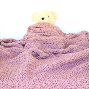 May include: A soft, pink crocheted blanket with a teddy bear tucked inside. The blanket is made with a delicate stitch pattern and has a cozy, comforting feel.
