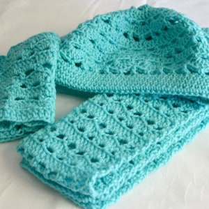 Puede incluir: Un conjunto turquesa de gorro, bufanda y guantes sin dedos de ganchillo. El conjunto hecho a mano presenta un diseño calado. El gorro está doblado y la bufanda está cuidadosamente colocada. Los guantes están colocados a la izquierda del gorro y la bufanda.