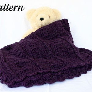 Può includere: Una coperta all'uncinetto di colore viola con un bordo a festoni, che presenta un motivo testurizzato. Un orsacchiotto di peluche è sdraiato sopra la coperta.