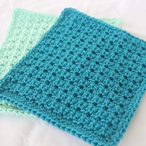 May include: Two handmade crochet washcloths. One is a vibrant teal, the other a soft mint green. The washcloths are square, with a textured stitch pattern. They are likely made of cotton yarn, suitable for the kitchen or bathroom.