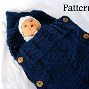 May include: A navy blue knitted baby sleeping bag with a hood and wooden buttons. The sleeping bag is made with a cable knit pattern.