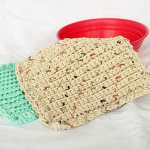 May include: Two crocheted dishcloths, one in a light green color and the other in a beige color with brown and orange flecks. The dishcloths are on a white surface.