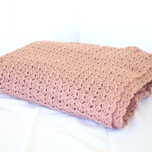 May include: A soft, pink crocheted blanket with a delicate scalloped edge. The blanket is folded and displayed on a white surface.