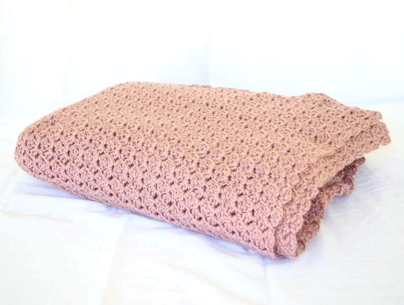 May include: A soft, pink crocheted blanket with a delicate scalloped edge. The blanket is folded and displayed on a white surface.
