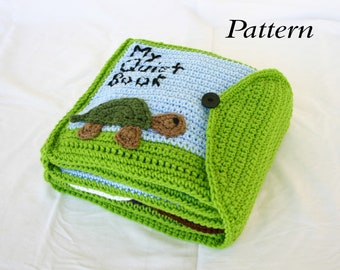 May include: A green crocheted baby book with a blue and white cover. The cover has the text "My First Book" and a green crocheted turtle with brown feet. The book has a green crocheted flap.