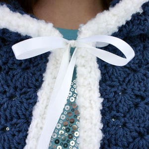 K&ouml;nnte beinhalten: Eine blaue H&auml;kel-Kapuzenjacke mit wei&szlig;em H&auml;kelrand und einem wei&szlig;en Satinband, das am Hals gebunden ist.
