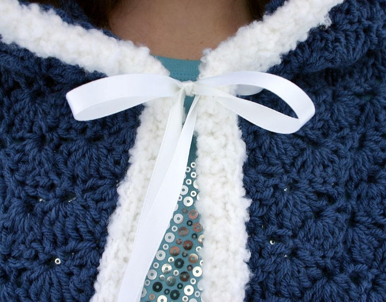 K&ouml;nnte beinhalten: Eine blaue H&auml;kel-Kapuzenjacke mit wei&szlig;em H&auml;kelrand und einem wei&szlig;en Satinband, das am Hals gebunden ist.