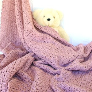 May include: A soft, pink crocheted blanket with a delicate stitch pattern. The blanket is draped over a white surface with a plush, cream-colored teddy bear peeking out from underneath.