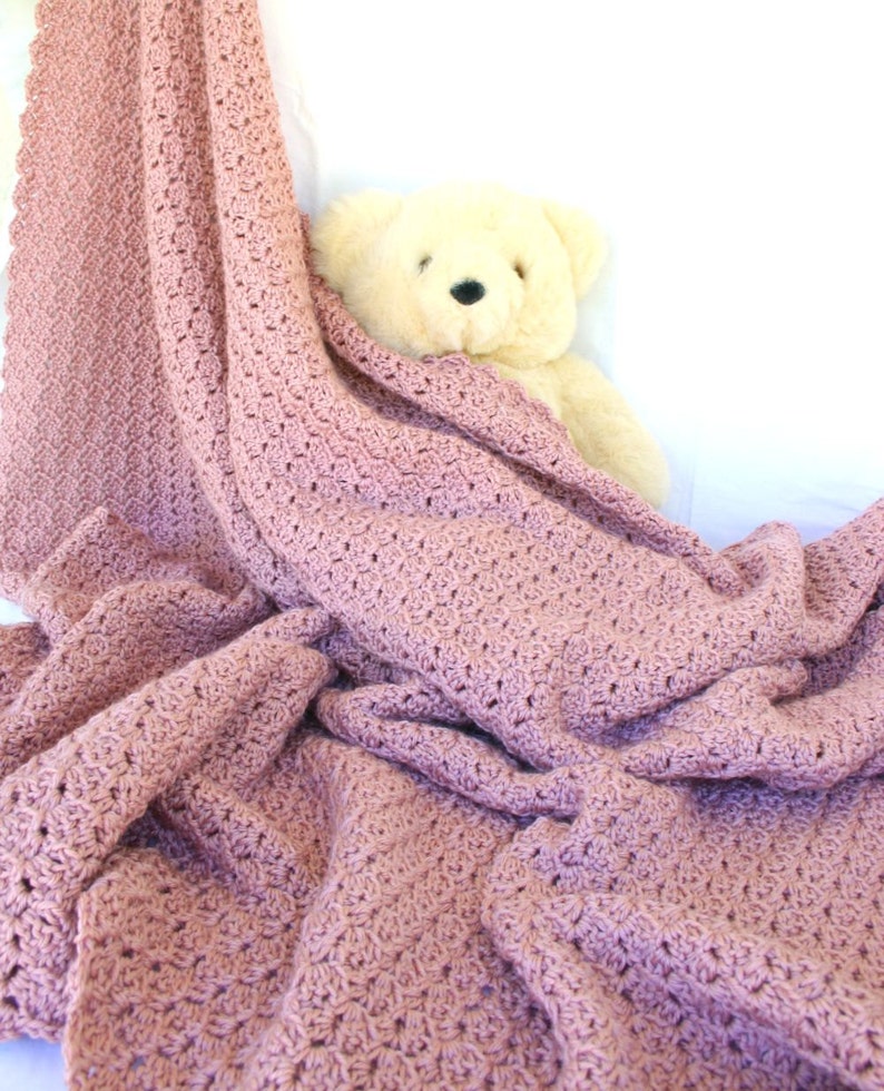 May include: A soft, pink crocheted blanket with a delicate stitch pattern. The blanket is draped over a white surface with a plush, cream-colored teddy bear peeking out from underneath.