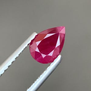 Op de afbeelding: Een peervormige, gefacetteerde robijn met een diepe, levendige rode kleur. De edelsteen wordt vastgehouden door een zilverkleurige metalen pincet, wat de heldere, gepolijste oppervlakken en de precieze geometrische slijpvorm benadrukt.