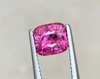 May include: A vibrant, cushion-cut, pink gemstone held by silver tweezers. The gemstone has multiple facets that reflect light, creating a sparkling effect. The color is a deep, saturated pink, and the overall shape is square with rounded corners. This gemstone is ideal for jewelry making.