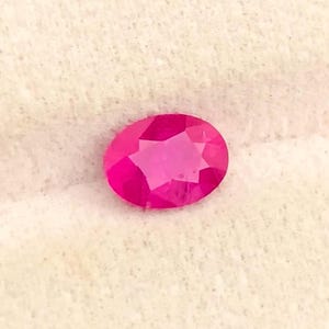 May include: A vibrant, oval-cut gemstone with a deep, saturated pinkish-red hue. The stone features a faceted surface that reflects light, creating a brilliant, sparkling appearance across its polished, symmetrical, and translucent structure.