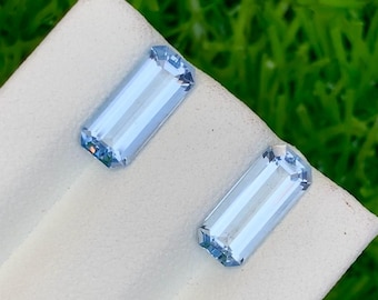 Op de afbeelding: Twee rechthoekige, lichtblauwe edelstenen met een facetgeslepen slijpsel worden getoond op een witte ondergrond tegen een achtergrond van groen gras. De edelstenen hebben een subtiele, ijzige uitstraling, die het licht reflecteert en hun helderheid toont.