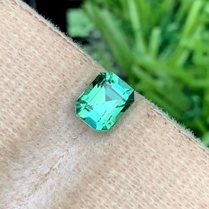 K&ouml;nnte beinhalten: Ein rechteckiger, smaragdgeschliffener Edelstein mit einem leuchtenden, hellgr&uuml;nen Farbton. Der Edelstein ist facettiert, reflektiert das Licht und zeigt seine Klarheit. Der Hintergrund ist eine weiche, neutrale Farbe, mit gr&uuml;nem Laub im Hintergrund.