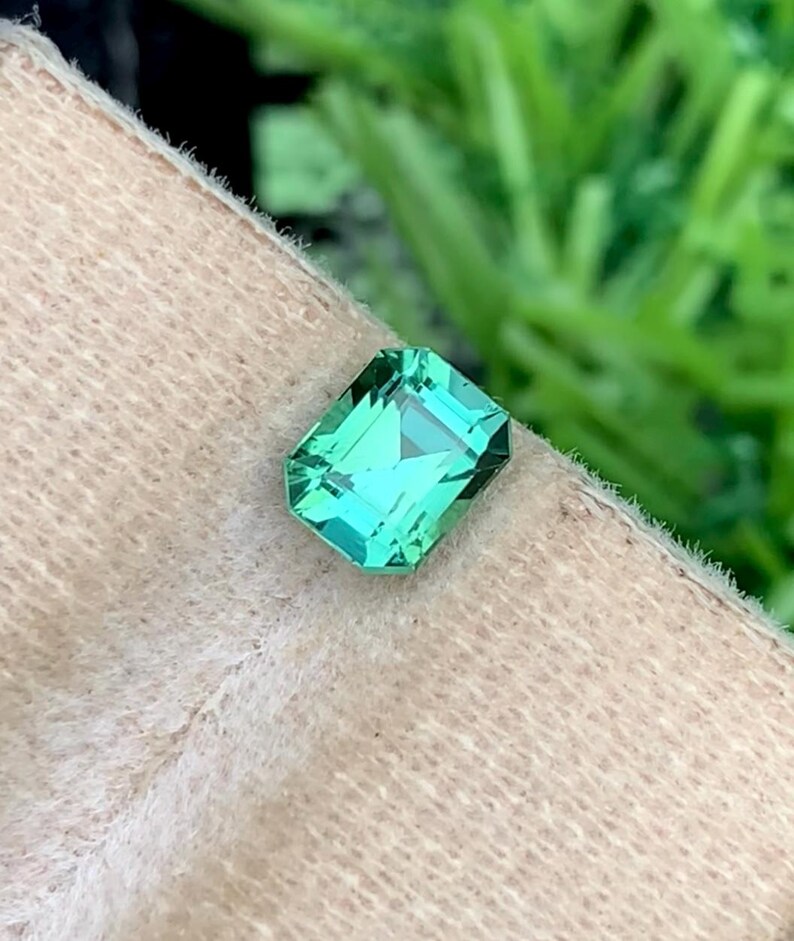 K&ouml;nnte beinhalten: Ein rechteckiger, smaragdgeschliffener Edelstein mit einem leuchtenden, hellgr&uuml;nen Farbton. Der Edelstein ist facettiert, reflektiert das Licht und zeigt seine Klarheit. Der Hintergrund ist eine weiche, neutrale Farbe, mit gr&uuml;nem Laub im Hintergrund.