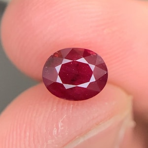 May include: An oval-cut ruby gemstone with a deep red color and a faceted surface. The stone displays a clear, symmetrical pattern of triangular and kite-shaped facets that reflect light, creating a brilliant appearance across the entire surface.