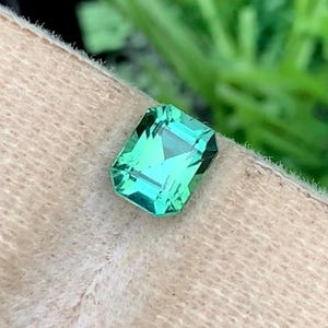 K&ouml;nnte beinhalten: Ein leuchtend hellgr&uuml;ner, smaragdgeschliffener Edelstein. Der Stein liegt vor einem neutralen, strukturierten Hintergrund, mit einem verschwommenen gr&uuml;nen Hintergrund. Die Facetten des Steins reflektieren das Licht und erzeugen einen funkelnden Effekt. Ideal f&uuml;r Schmuck.