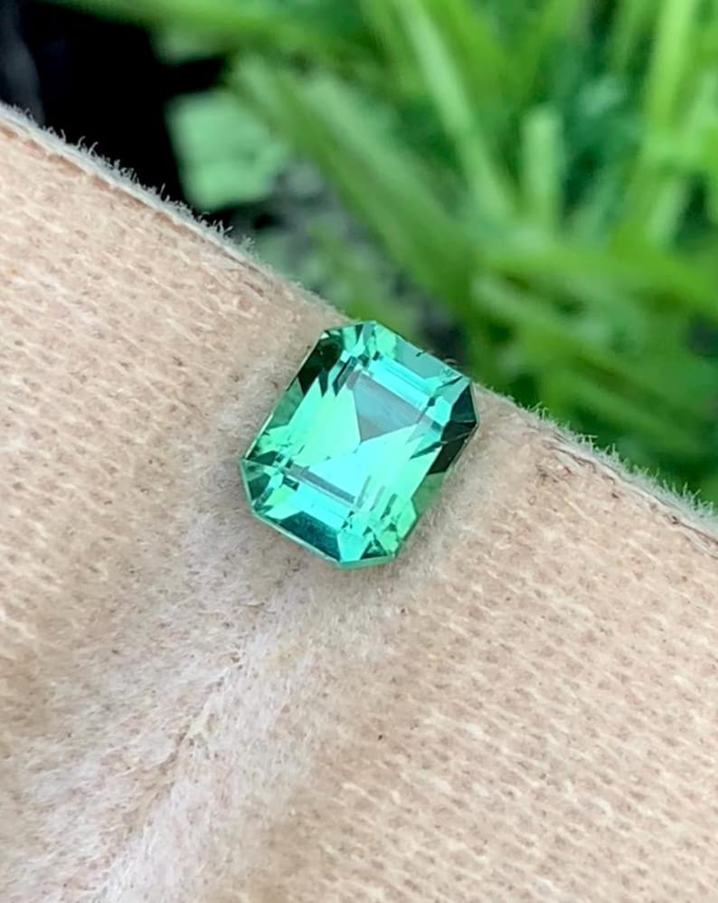 K&ouml;nnte beinhalten: Ein leuchtend hellgr&uuml;ner, smaragdgeschliffener Edelstein. Der Stein liegt vor einem neutralen, strukturierten Hintergrund, mit einem verschwommenen gr&uuml;nen Hintergrund. Die Facetten des Steins reflektieren das Licht und erzeugen einen funkelnden Effekt. Ideal f&uuml;r Schmuck.