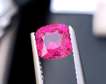 May include: A vibrant, cushion-cut, hot pink gemstone is held by silver tweezers. The gem has a faceted surface that reflects light, showcasing its clarity and color. The background is a blurred dark gray.