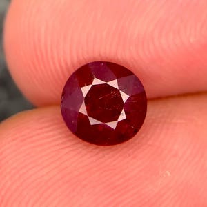 May include: A round, faceted, deep red gemstone with a brilliant cut. The stone displays a vibrant crimson hue and a symmetrical arrangement of triangular and kite-shaped facets that reflect light across its surface, creating a clear star pattern.