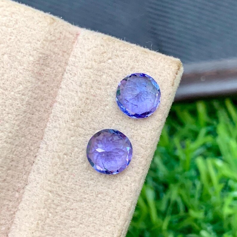 May include: Two round, faceted, purple gemstones on a beige background. The gemstones are approximately 1/2 inch in diameter.