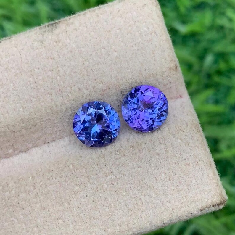 May include: Two round, faceted, purple and blue gemstones on a beige background. The gemstones are approximately 1 cm in diameter.