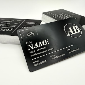 May include: A stack of rectangular black business cards with white text. The front card features a circular AB logo, a placeholder for a logo, and fields for full name, title, position, tagline, phone number, email, and website in a clean layout.