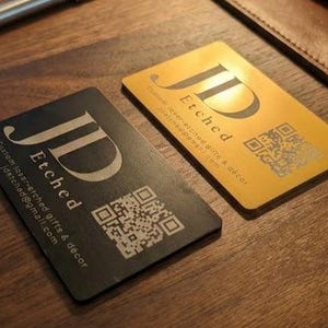 May include: Two rectangular business cards, one black and one gold, with the letters "JD" and the word "Etched" in a stylized font. Each card has a QR code and contact information. The cards are on a wooden surface.