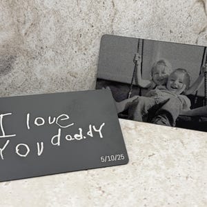 May include: Two rectangular metal wallet cards with rounded corners. One card features a black and white engraved photograph of two children on a swing. The other card is dark gray with the white engraved text I love you daddy and 5/10/25.