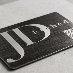May include: A rectangular metal business card with the etched text "JD Etched" and a QR code. The card has a brushed metal texture and is displayed on a light gray surface. The design is in a minimalist style.