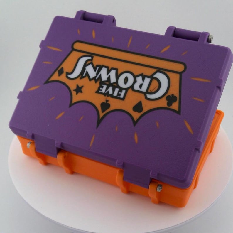 May include: A purple and orange plastic case with the words "FIVE CROWNS" in white lettering. The case has a hinged lid and a clasp closure. The top of the case has a graphic design with a star and card suit symbols.