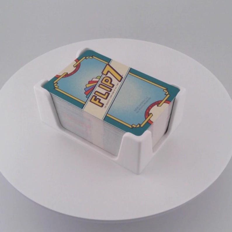 May include: A white plastic card holder containing a deck of cards. The cards have a blue and gold design with the text "FLIP 7" on the top card. The holder is on a white surface.